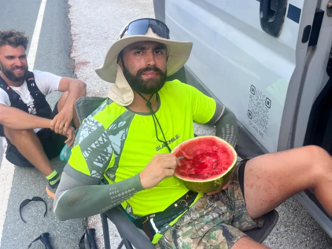 The athlete sitting on the ground against the van eating watermelon