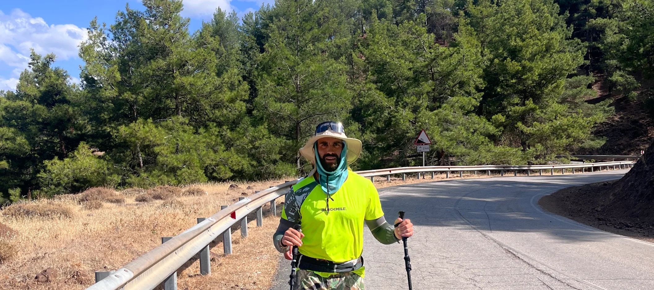 Andreas Nikolaou on a pine-lined mountain road with trekking poles, in a wide-brimmed hat and neck gaiter, in bright afternoon sun