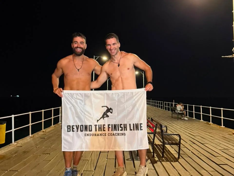Two men holding a Beyond the Finish Line Endurance Coaching banner on the Molos Pier boardwalk at night